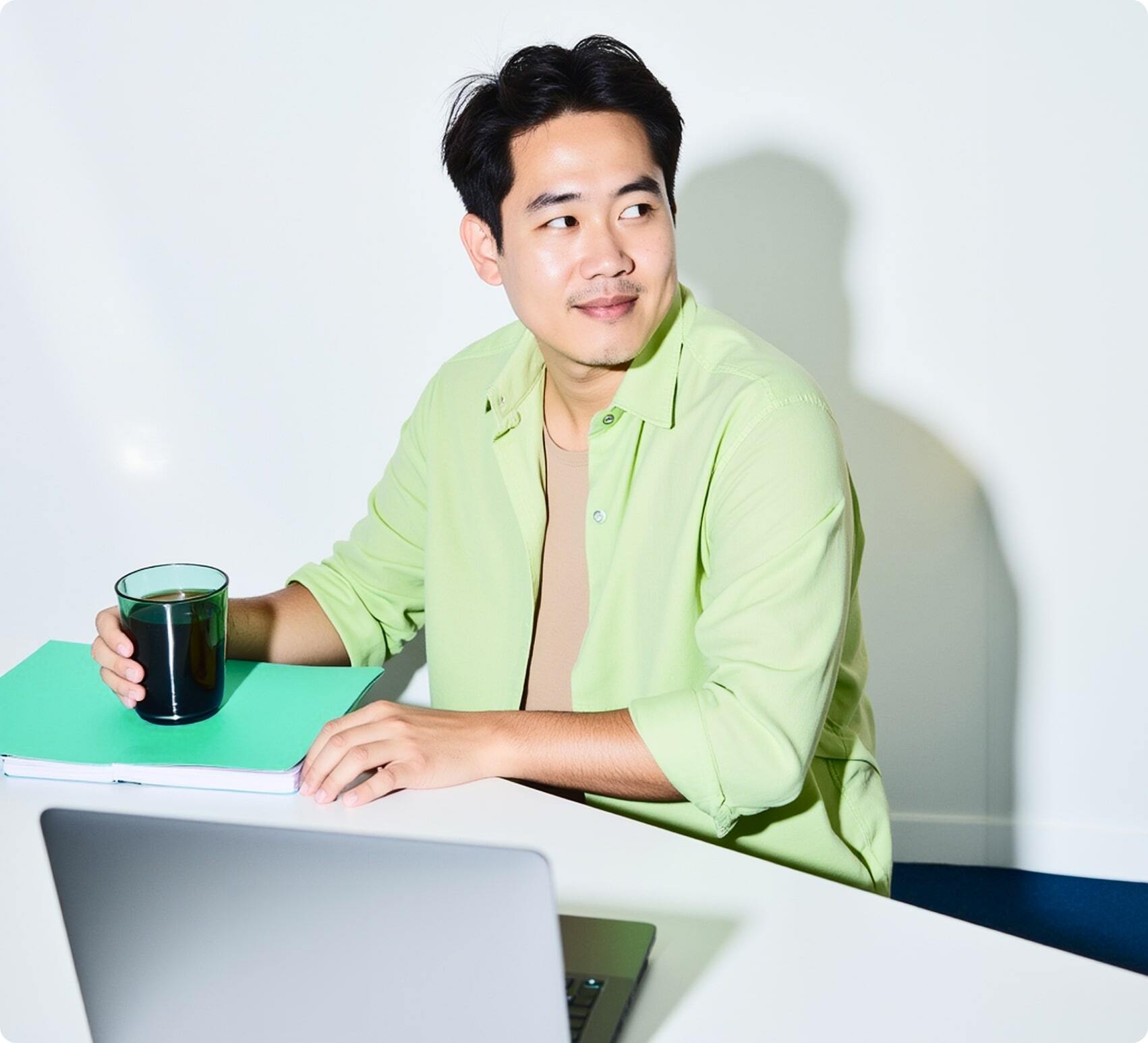 Man in a light green shirt sitting at a desk with a laptop, holding a glass with a dark drink, looking to the side and smiling.